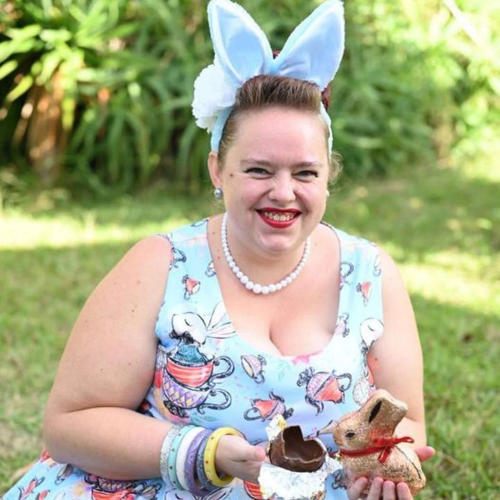 Woman in blue dress with rabbit print, wearing bunny ears, holding chocolate bunny in a garden setting.