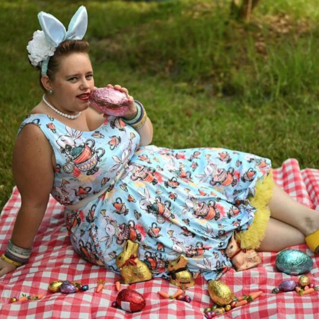 Woman in whimsical skater dress with bunny ears, enjoying Easter treats on a picnic blanket.