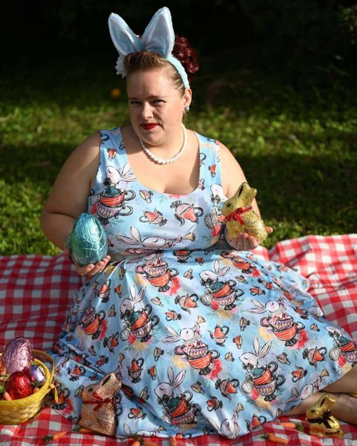 Woman in a whimsical blue skater dress with bunny prints, seated on a picnic blanket, surrounded by Easter eggs and decorations.